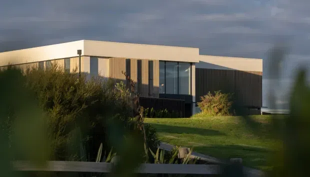 Exterior view of a low-slung contemporary single-storey house with a flat roof, dark vertical cladding, pale rendered walls, and large glazed openings. The building is set within landscaped gardens and photographed in warm, low evening light.