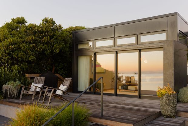 Exterior view of a compact contemporary pavilion or extension with a flat roof, dark grey framed glazing, and a concrete-like facade. It opens onto a timber deck with outdoor lounge chairs, planting, and a simple dark metal handrail. Warm interior light and expansive glazing reflect a coastal or waterfront outlook, though the distant view is secondary.