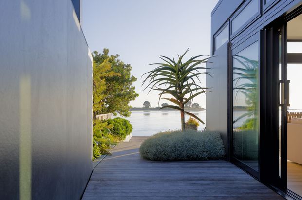 A contemporary waterfront residential exterior viewed from a timber deck or narrow outdoor passage. A dark, smooth-clad wall runs along the left, while the right side has large black-framed glazed folding or sliding doors opening into a bright interior. The deck leads toward calm water, with landscaped planting including a sculptural palm-like tree and rounded shrubs. The scene has clean modern lines, strong indoor–outdoor connection, and tranquil coastal character.