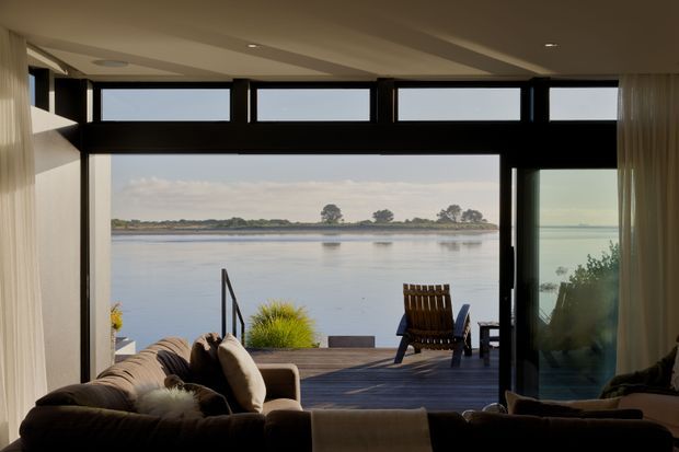 A calm waterfront living room opening onto a timber deck, with expansive black-framed glazing and clerestory windows overlooking a broad body of water. The room contains a large upholstered sofa, lounge seating, sheer curtains, and a timber outdoor chair, creating a relaxed indoor–outdoor connection.