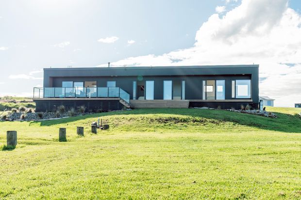 A low, elongated contemporary house set on a gently sloping grassy site. The dark horizontal facade has extensive floor-to-ceiling glazing, a flat roof, a raised timber-clad central entry or terrace section, and a deck with glass balustrading. Bright open sky and landscaped lawn surround the building.