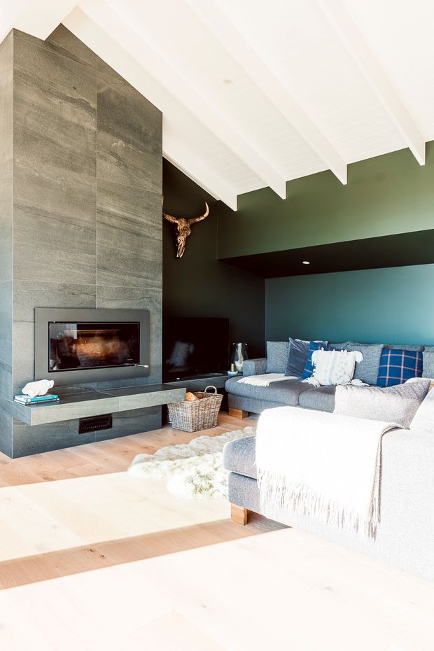 Bright, cozy living room with a large grey sectional sofa arranged around a contemporary built-in fireplace. The room features a dramatic dark green and black accent wall, expansive grey stone fireplace surround and hearth, pale timber flooring, white painted timber-lined ceiling with exposed beams, and layered blue and white cushions and throws. A decorative bull skull is mounted above the fireplace area.