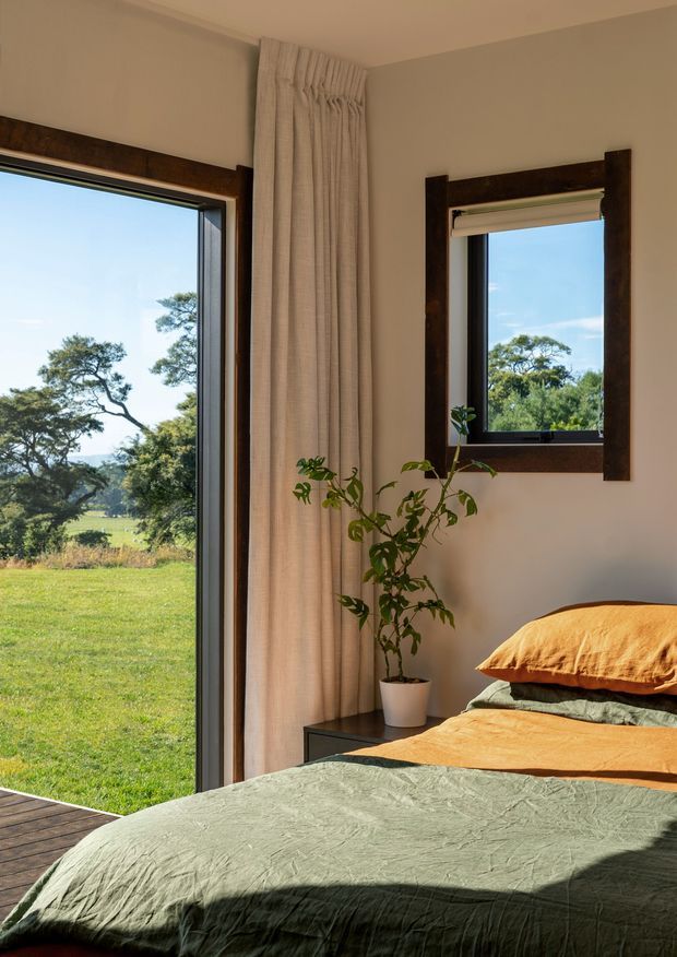 A sunlit bedroom with a low bed dressed in textured sage-green and ochre bedding. Full-height glazing opens toward a grassy rural landscape, while a smaller framed window, linen curtains, a potted plant, and dark timber window trim add warmth and natural character.
