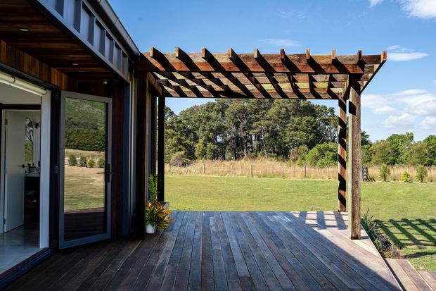 Exterior view of a contemporary dark-clad building opening onto a large timber deck and covered pergola. The pergola has exposed timber beams and rafters, with glass or glazed doors connecting to the interior. The deck overlooks a grassy rural landscape and mature trees under a clear sky.