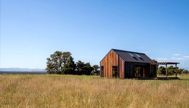 A small rural cabin or barn-like dwelling set in a wide grassland field. The building has dark vertical timber cladding, a steep simple gable roof with dark metal roofing, and an attached open-sided covered deck or verandah. Mature trees and distant hills form the background beneath a clear blue sky.