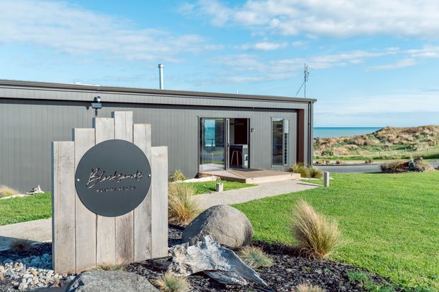 A low, single-storey contemporary building with dark grey vertical cladding, a shallow flat or low-pitched roof, and glazed entry doors. A timber monument sign stands in the landscaped foreground, with ornamental grasses, rocks, lawn, and a visible coastal horizon beyond. The image appears to show a commercial or visitor-oriented property exterior in a coastal setting.