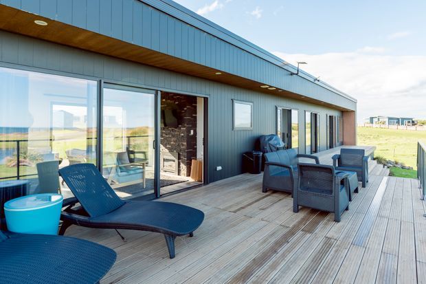 A contemporary single-storey home viewed from a spacious timber deck. The exterior has dark vertical metal cladding, large sliding glazed doors and windows, and a warm timber-lined soffit. Outdoor lounge furniture and sun loungers occupy the deck, with open rural fields visible beyond.