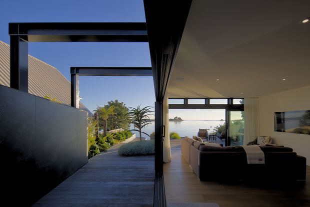 A contemporary indoor-outdoor living space viewed along a covered passage toward a waterfront outlook. The image shows a minimalist living room with large glazed openings, dark-framed windows and doors, pale flooring, neutral walls, and dark upholstered sofas. A landscaped path and garden sit to the left, while the living area opens toward the water and a small outdoor seating area.