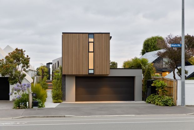 Street-facing view of a compact contemporary multi-storey residence with a dark double garage at ground level, a projecting upper volume clad in vertical timber battens, and a narrow vertical strip of windows. The home sits within a landscaped suburban streetscape with adjacent residential properties and fencing visible.