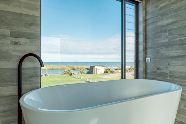 A modern bathroom featuring a large freestanding white bathtub positioned beside full-height glazing, with a dark curved floor-mounted tap. The walls are clad in grey wood-look panels, and the view through the window overlooks a lawn, coastal dunes, and the ocean. The space is bright, refined, and strongly connected to the coastal setting.