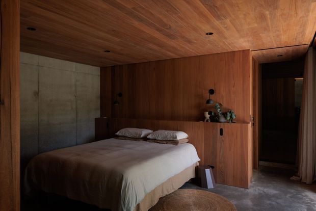 A moody, compact bedroom with a low-profile bed set into extensive dark timber wall panelling and built-in joinery. The timber continues across the ceiling, creating a warm enveloping effect, while a raw concrete feature wall contrasts on the left. Recessed downlights, a small open shelf with plants, and a dark ensuite or wardrobe opening are visible at the rear. The room has a soft neutral bed covering and a textured round rug on a polished grey floor.