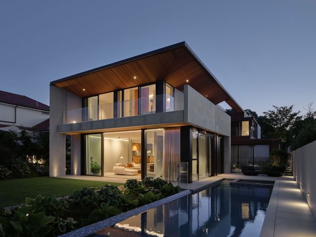 Contemporary two-storey residence photographed at dusk, with strong horizontal roof planes, extensive floor-to-ceiling glazing, a deep timber-lined roof soffit, and smooth grey rendered or concrete-like walls. The house opens onto a narrow rectangular swimming pool bordered by paved paths and landscaped garden beds. Warm interior lighting and restrained modern detailing create a refined indoor-outdoor setting.