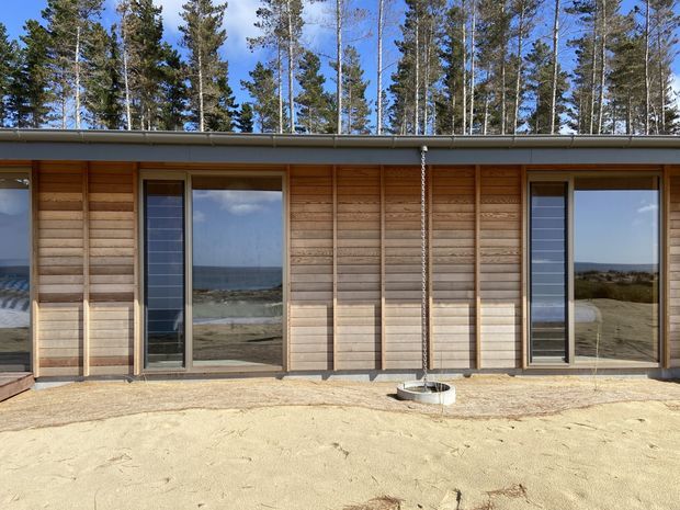 Single-storey modern coastal building facade with horizontal timber cladding, tall glazed doors and windows, and a low-profile metal roof. The reflective glazing shows a sandy coastal landscape and ocean beyond, while tall pine trees rise behind the structure.