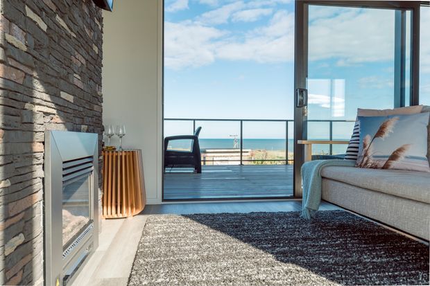 A bright coastal living room with a full-height glazed sliding door opening onto a balcony or terrace overlooking the ocean. The room features a substantial natural stacked-stone fireplace wall, pale timber flooring, a textured grey rug, a light upholstered sofa with striped and feather-patterned cushions, and a warm timber console. A dark metal balcony balustrade and outdoor chair are visible beyond the glazing.