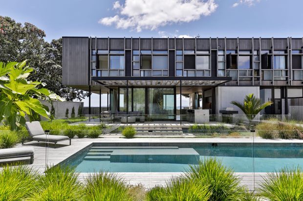 A contemporary multi-storey residence viewed from the landscaped pool area. The dark, vertically ribbed facade has a strong rectilinear grid of projecting frames and large windows, with a glazed ground-floor living area beneath a deep cantilevered upper level. A rectangular swimming pool, paved terrace, outdoor lounge furniture, ornamental grasses and tropical planting occupy the foreground.