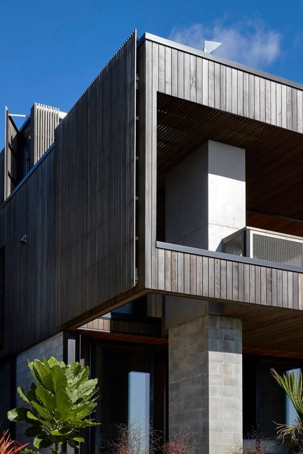 A close-up exterior view of a contemporary multi-level building with dramatic cantilevered forms, vertical dark timber cladding, exposed concrete block supports, and a recessed balcony or outdoor opening. The balcony soffit is lined with timber slats, and perforated metal balustrading is visible. Strong sunlight creates sharp shadows against a clear blue sky, with limited landscaping at the base.