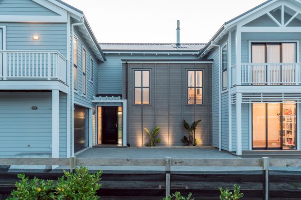 Exterior view of a coastal-style multi-level residential building arranged around a central courtyard. The facade features pale blue horizontal weatherboards, white trim, gabled roof forms, upper-level balconies with balustrades, and large glazed doors and windows. A dark grey weatherboard central entry volume is illuminated with wall lights and small planted areas. A timber retaining or boundary structure occupies the foreground.