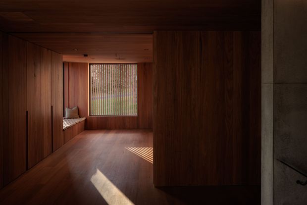 A dark, richly timber-lined interior corridor or compact living space, with extensive built-in wood cabinetry along both sides, timber flooring and ceiling, and a built-in window seat at the far end. Vertical blinds or slatted screening filter daylight through the window, casting narrow bands of sunlight across the floor. A partial concrete wall with a simple dark handrail is visible at right. The composition is minimalist, warm, and highly integrated, with no loose furnishings beyond cushions on the bench seat.
