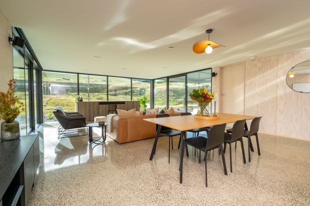 A bright, open-plan contemporary living and dining area with extensive floor-to-ceiling glazing overlooking a rural landscape. The room features a tan sectional sofa, black leather lounge chair, timber dining table with black chairs, a polished terrazzo floor, dark-framed windows and doors, and a pale timber-panelled feature wall. Sculptural pendant lighting, indoor plants and warm furnishings create a refined, inviting interior.