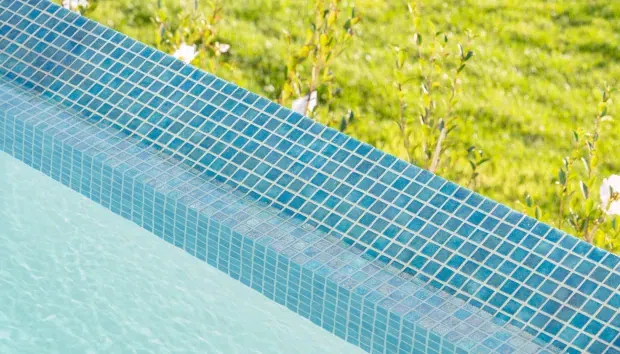 Close-up detail of a swimming pool edge lined with small square turquoise-blue mosaic tiles. The curved coping and waterline are visible, with a blurred green lawn and planting beyond the pool.