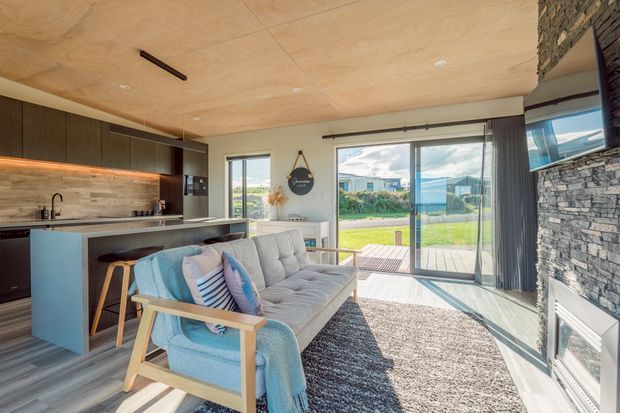 Bright open-plan living area with a compact kitchen, timber cabinetry, island bench, and a mid-century-style sofa facing a stone-clad fireplace wall. Large sliding glass doors open to a lawn and outdoor deck, while a plywood-lined ceiling and warm timber finishes create a contemporary, natural interior.