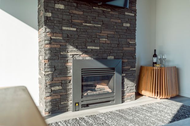 Bright contemporary living room featuring a full-height stacked-stone fireplace surround with an inset gas fire. A sculptural ribbed timber sideboard with wine and glassware sits to the right, while a textured grey rug and pale walls complete the warm, refined setting.