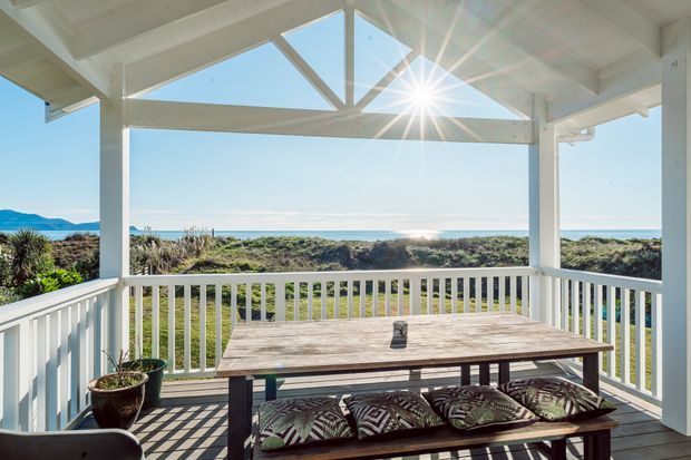 A bright covered coastal deck or veranda with white-painted timber posts, railings and exposed triangular roof framing. A long weathered timber picnic table with patterned cushions occupies the foreground, while low coastal vegetation, sandy dunes and the ocean are visible beyond. Strong sunlight and expansive sea views create an airy outdoor entertaining setting.