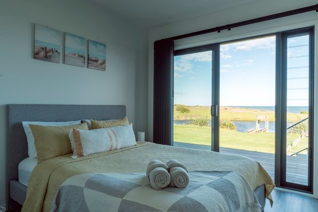A bright coastal bedroom with a neatly made bed positioned beside expansive dark-framed sliding glass doors. The room has a pale blue-grey upholstered headboard, neutral and muted blue bedding, minimal wall art, and a direct view toward a sandy coastal landscape and ocean beyond. The overall atmosphere is calm, airy, and relaxed.