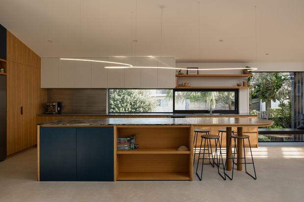 Contemporary open-plan kitchen with a large island featuring dark cabinetry, warm timber open shelving, and a stone benchtop. White overhead cabinets and integrated linear lighting create a clean, streamlined appearance. A timber wall of tall cabinetry, rear workbench, bar seating, and extensive glazing to a leafy outdoor area are visible.