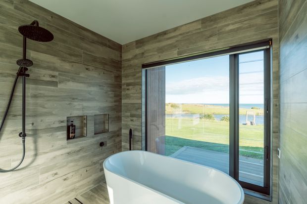 A modern bathroom featuring a freestanding white bathtub beside full-height glazing with ocean views. The walls and floor are clad in wood-look grey-brown tiles, with a dark bronze or black rainfall shower, recessed niches, and a large sliding glass door opening toward an exterior deck or lawn.