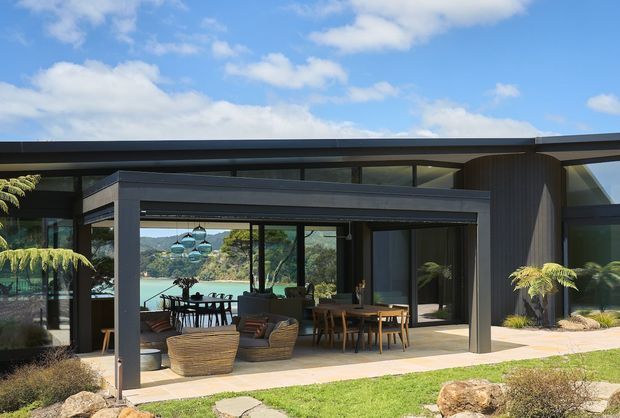 Contemporary single-storey residence with a dark, low-profile flat roof and extensive floor-to-ceiling glazing. The image focuses on a covered outdoor entertaining terrace furnished with lounge seating and a dining table, opening toward a turquoise waterfront view. Dark vertical cladding, slim black window frames, and a landscaped garden reinforce the modern indoor-outdoor design.
