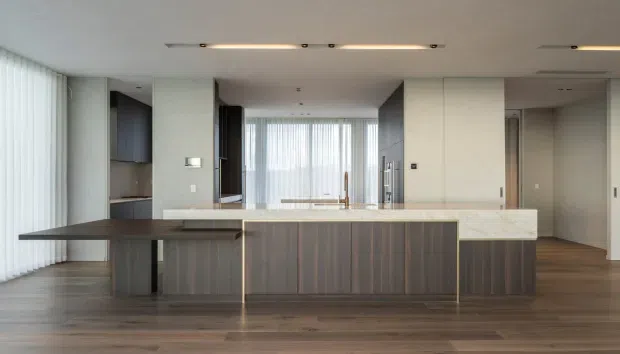A spacious contemporary kitchen with a large central island, dark timber cabinetry, a pale stone benchtop, integrated appliances, and a projecting dark breakfast bar. Floor-to-ceiling sheer glazing lines the left side, while slim recessed linear lights and warm timber flooring create a clean, refined interior.