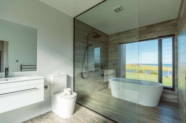Bright contemporary bathroom with a floating white vanity, wall-mounted mirror, toilet, walk-in glass shower, and freestanding white bathtub. Large windows provide expansive views toward a blue waterway and open landscape. Wood-look wall and floor finishes add warmth to the otherwise clean, modern space.