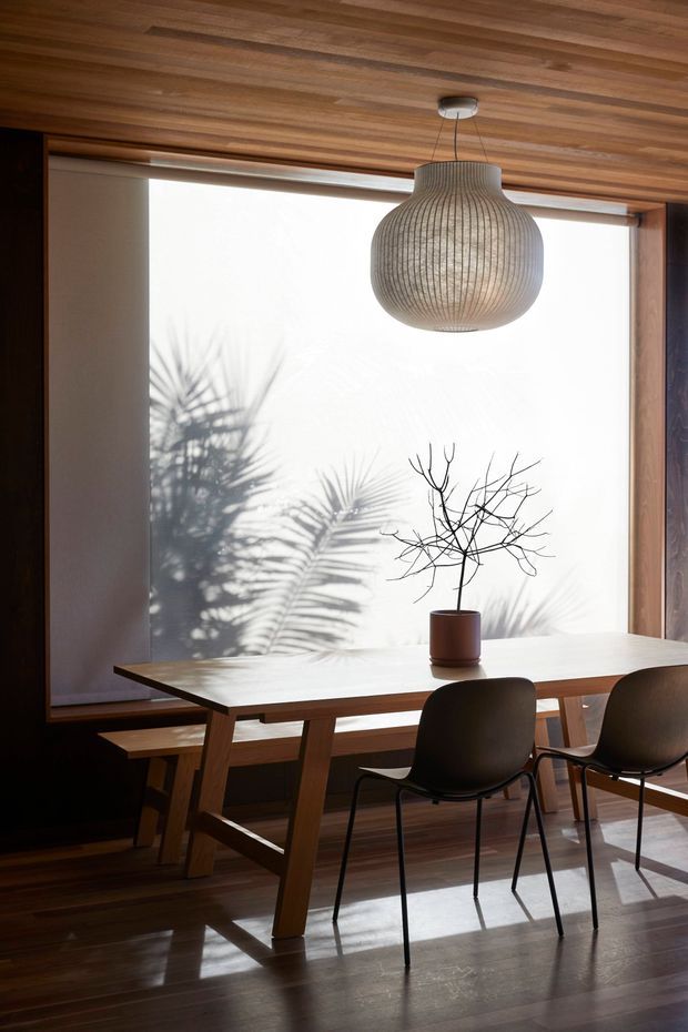 A warm, minimalist dining area with a solid timber dining table and dark chairs set against a large bright window. The room features extensive timber lining on the ceiling and walls, polished dark timber flooring, a sculptural pendant light, and a simple potted branch arrangement. Strong palm-leaf shadows create a calm, natural atmosphere.