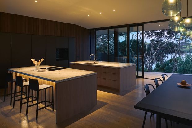 A sophisticated open-plan kitchen and dining area with two large stone-topped islands, dark timber cabinetry and wall panelling, integrated appliances, and black bar and dining furniture. Full-height glazed doors and windows open the room to a leafy outdoor setting. Warm recessed lighting and clustered glass pendant lights create an atmospheric, refined interior.