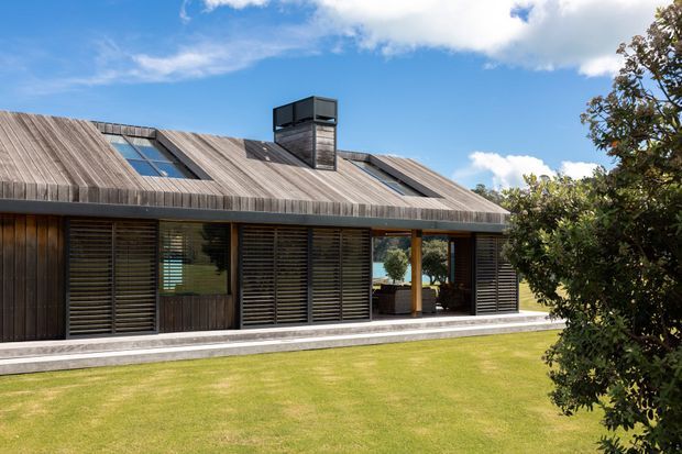 Exterior view of a low, modern single-storey house with a broad pitched roof clad in weathered timber, integrated skylights, and a central roof vent or chimney structure. The facade combines dark timber cladding with extensive horizontal louvred glazing and sheltered outdoor living areas. A manicured lawn and mature planting surround the building under a bright blue sky.