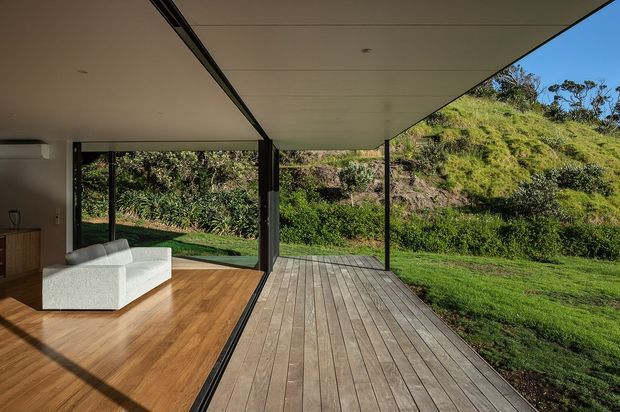 A modern indoor-outdoor living area with full-height sliding glass openings connecting a timber-floored interior lounge to a large timber deck. Slim dark-framed glazing and slender structural posts support a broad flat roof overhang, while landscaped greenery and a sloping garden are visible beyond. The space is sparsely furnished with a white sofa and has strong indoor-outdoor flow.