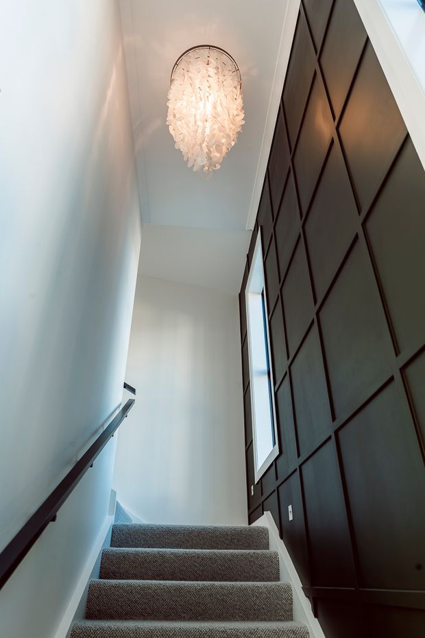 A narrow, tall interior stairwell viewed looking upward. The staircase has grey carpeted treads, white walls, a dark timber handrail, and a dramatic full-height dark timber feature wall with geometric panel moulding. A tall narrow window brings in daylight, while a sculptural white pendant light hangs from the ceiling.