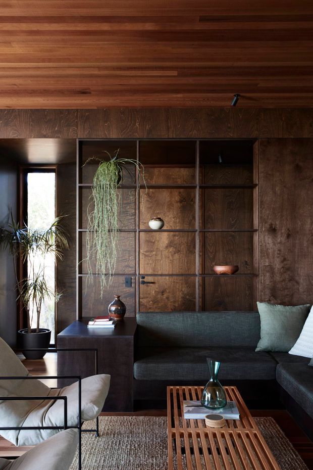 A richly layered living room with dark timber wall panelling, a matching timber ceiling, and custom open shelving integrated around a concealed door. A deep charcoal sectional sofa, light upholstered lounge chair, indoor plants, sculptural ceramics, and a slatted timber coffee table create a warm, refined and moody interior.