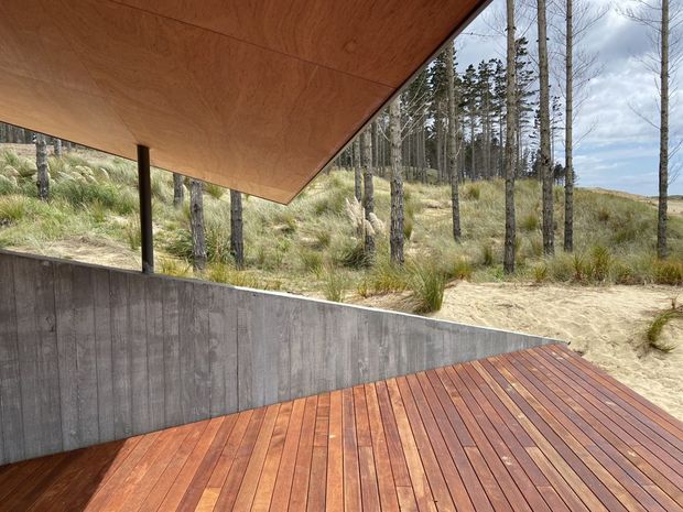 A minimalist timber-clad outdoor terrace or deck beneath a large angled timber canopy. The deck is bordered by a raw concrete retaining wall and opens toward a natural sandy landscape with grasses and tall trees. The image emphasises the strong geometric roof plane, warm timber surfaces, and seamless connection to the surrounding site.