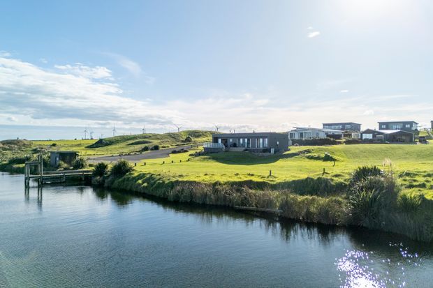 A wide exterior landscape view of a contemporary residential development beside a calm waterway. Several low-profile modern homes sit on gently rolling green grass, with a small waterside structure and dock at the left. The scene has expansive sky, bright sunlight, and a quiet coastal or rural character.