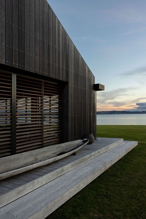 Exterior architectural view of a dark timber-clad pavilion or house beside a calm body of water. The facade features vertical blackened timber boards with visible fixings, large louvred screening over glazing, and a projecting box-like element. Broad pale timber decking and stepped platforms lead across a manicured lawn, with a weathered sculptural log or bench in the foreground. The scene is photographed at dusk with a muted sky and distant shoreline.
