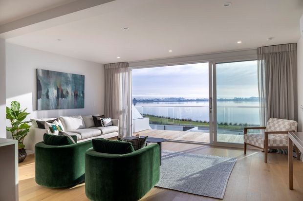 A bright, contemporary living room with a waterfront outlook through expansive floor-to-ceiling sliding glass doors. The room features pale timber flooring, white walls and ceiling, sheer curtains, a large neutral rug, a light sofa, deep green curved lounge chairs, and a patterned armchair. A spacious outdoor terrace and glass balustrade are visible beyond the glazing.