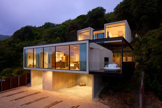 A modern multi-level hillside house at dusk, composed of cantilevered rectangular volumes with extensive floor-to-ceiling glazing, dark metal framing, and vertical timber-clad walls. Warm interior lighting reveals an open living and dining area, while an upper terrace includes outdoor seating. The lower level appears to contain an illuminated garage or covered entry, and dense vegetation surrounds the elevated structure.