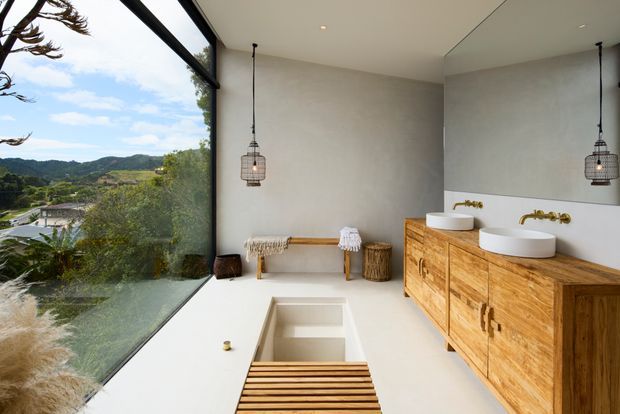 Contemporary minimalist bathroom with a deep freestanding soaking tub, dual vessel basins, wall-mounted brass taps, and a large timber vanity. Floor-to-ceiling glazing provides expansive views of a green hilly landscape, while pendant lights, a small bench, and a slatted timber bath platform add warmth and texture.