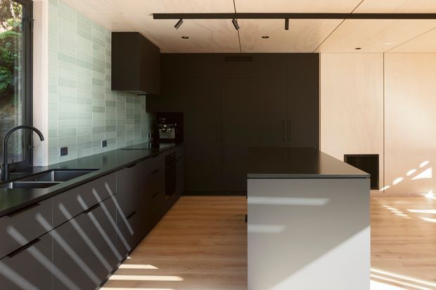 A contemporary minimalist kitchen with dark charcoal cabinetry along the left and rear walls, a central island, pale timber flooring, and a light wood-paneled wall. The kitchen includes a black sink and tap, dark benchtops, integrated appliances, a pale green tiled splashback, and a ceiling-mounted black linear track light. Strong sunlight creates diagonal shadows across the cabinetry, floor, and island.