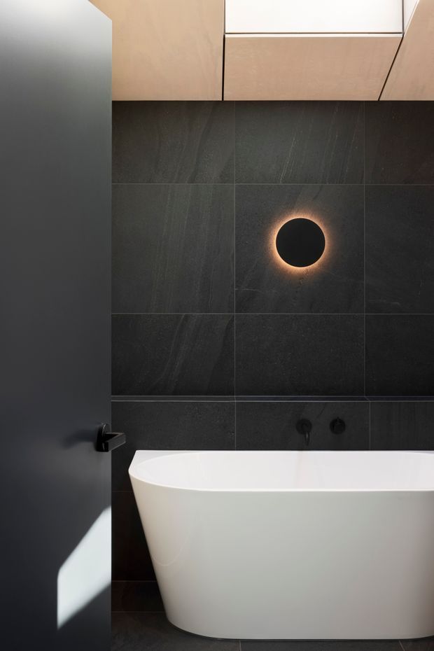 A compact, refined bathroom featuring a freestanding white bathtub against large dark charcoal stone-look wall tiles. A circular black wall detail with a warm halo light acts as a focal point, while pale wood-toned overhead cabinetry and a matte black door and tapware add contrast. The space has a dramatic, minimalist contemporary feel.