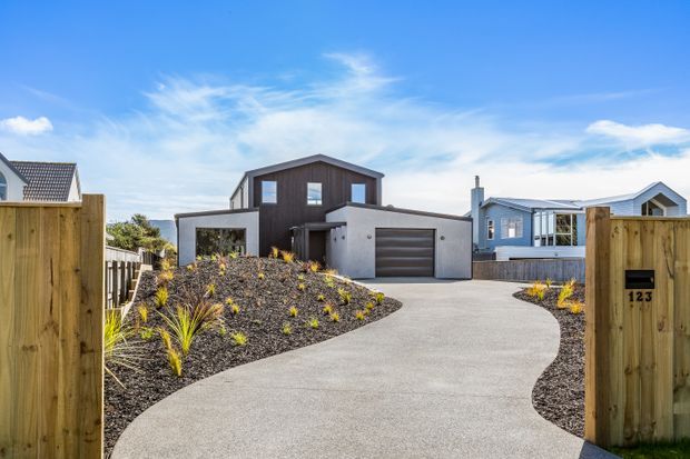 A contemporary two-storey home viewed from the street through a timber entrance gate. The dark vertical-clad upper volume sits above a light-rendered ground floor and integrated garage, with a curved concrete driveway and low-maintenance landscaped garden featuring ornamental grasses and yellow flowering plants. A neighbouring weatherboard home is visible to the side under a bright blue sky.
