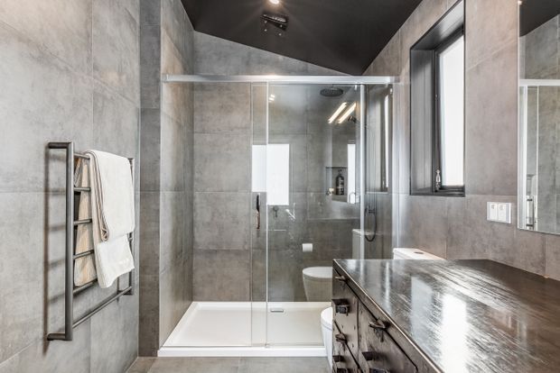 Contemporary bathroom featuring large-format grey concrete-look wall tiles, a glass-enclosed shower over a white bathtub, a dark timber vanity with integrated basin, wall mirror, black-framed window, and a heated towel rail. The dark ceiling and charcoal fixtures create a refined industrial feel.
