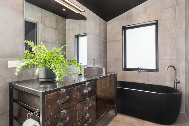A moody contemporary bathroom featuring a freestanding matte black bathtub, dark industrial-style metal vanity with drawers, concrete-look wall and floor tiles, black ceiling, black-framed windows, and a vessel basin. A green fern provides a natural accent.
